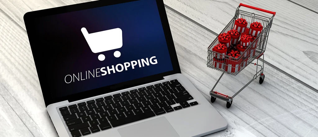 A laptop displaying an "Online Shopping" sign next to a miniature shopping trolley filled with wrapped gifts, symbolizing e-commerce and holiday shopping.