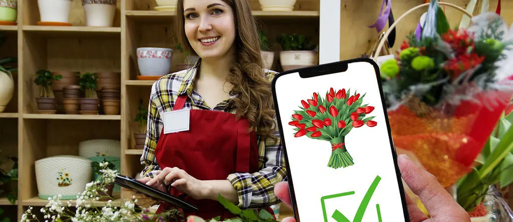 A person using a mobile phone to purchase flowers online, with a vibrant florist shop in the background displaying colorful blooms and arrangements.