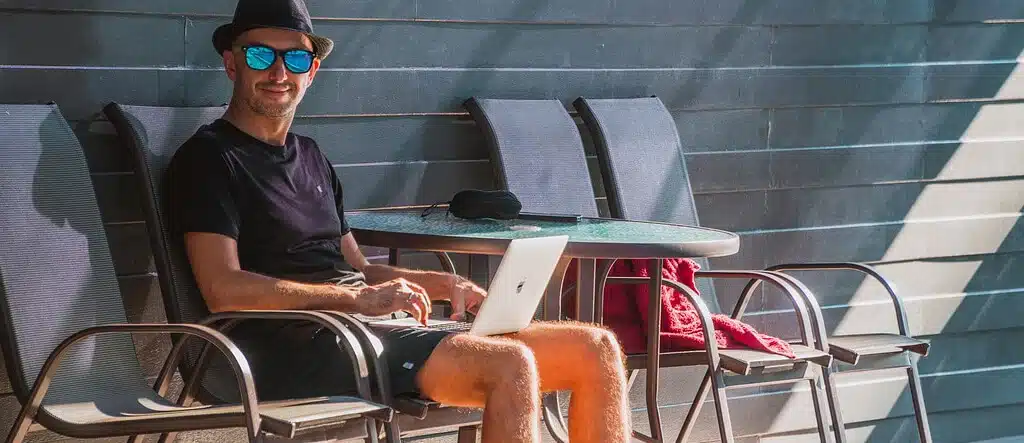 A man sitting on a comfy chair beside a pool, using his laptop, representing the flexibility and freedom of working from home or remotely