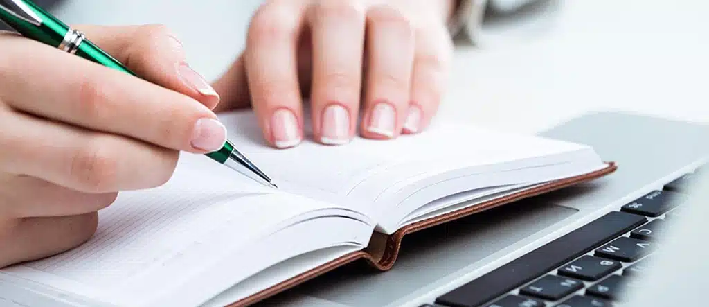 A person writing in a notebook placed on a laptop, symbolizing freelancing opportunities like content writing and working from home.