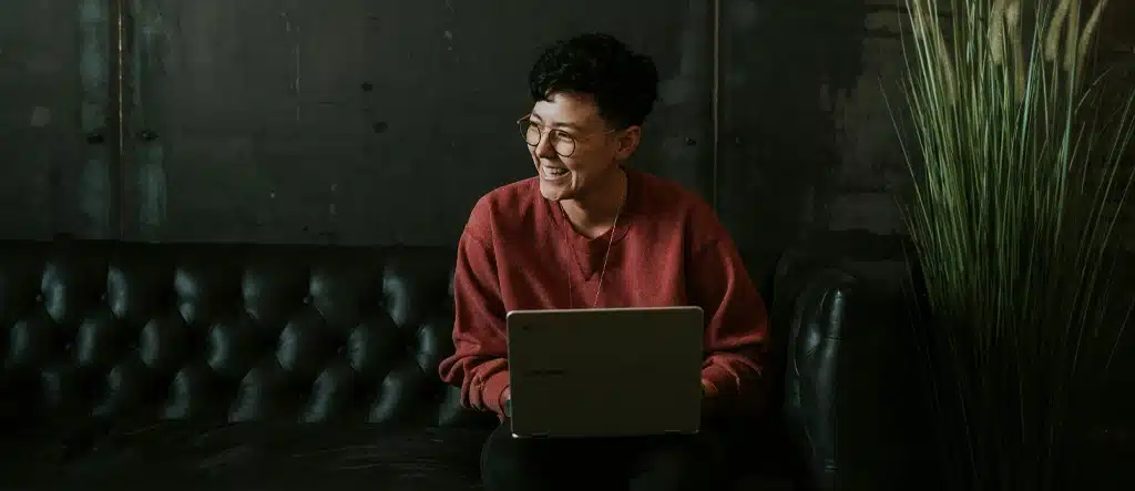 A woman sitting on a sofa with a laptop on her legs, smiling as she works on her home-based business.